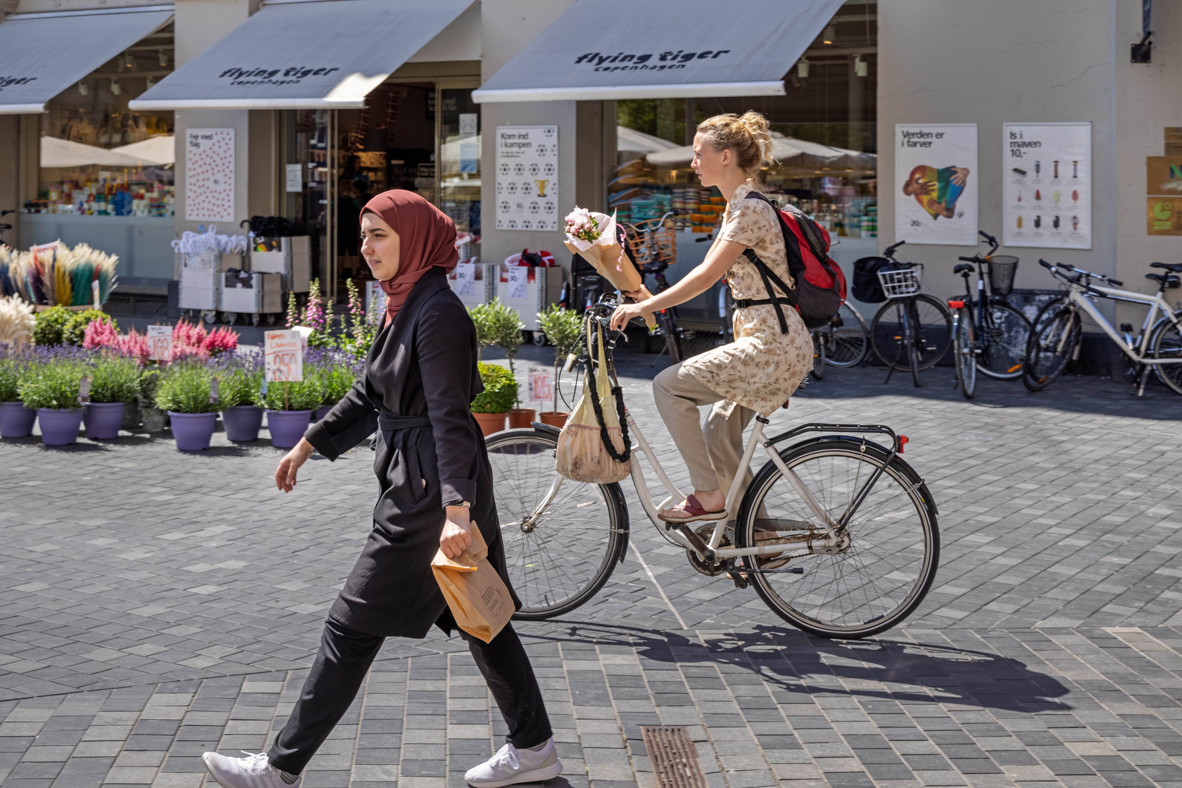 A woman walks through a square with a cyclist behind her riding along and holding a bunch of flowers