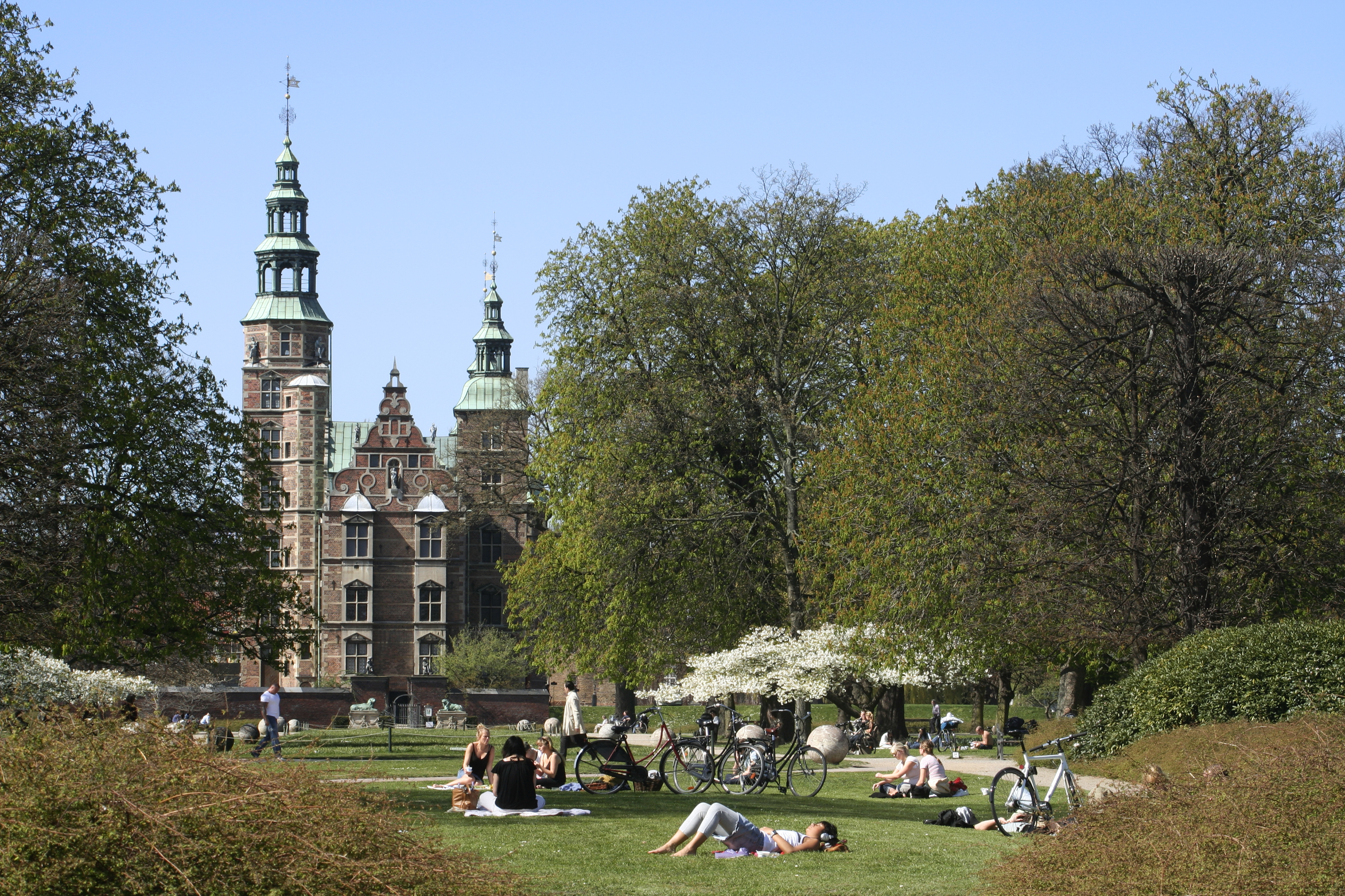 People relaxing in a park in front of a building with two towers 