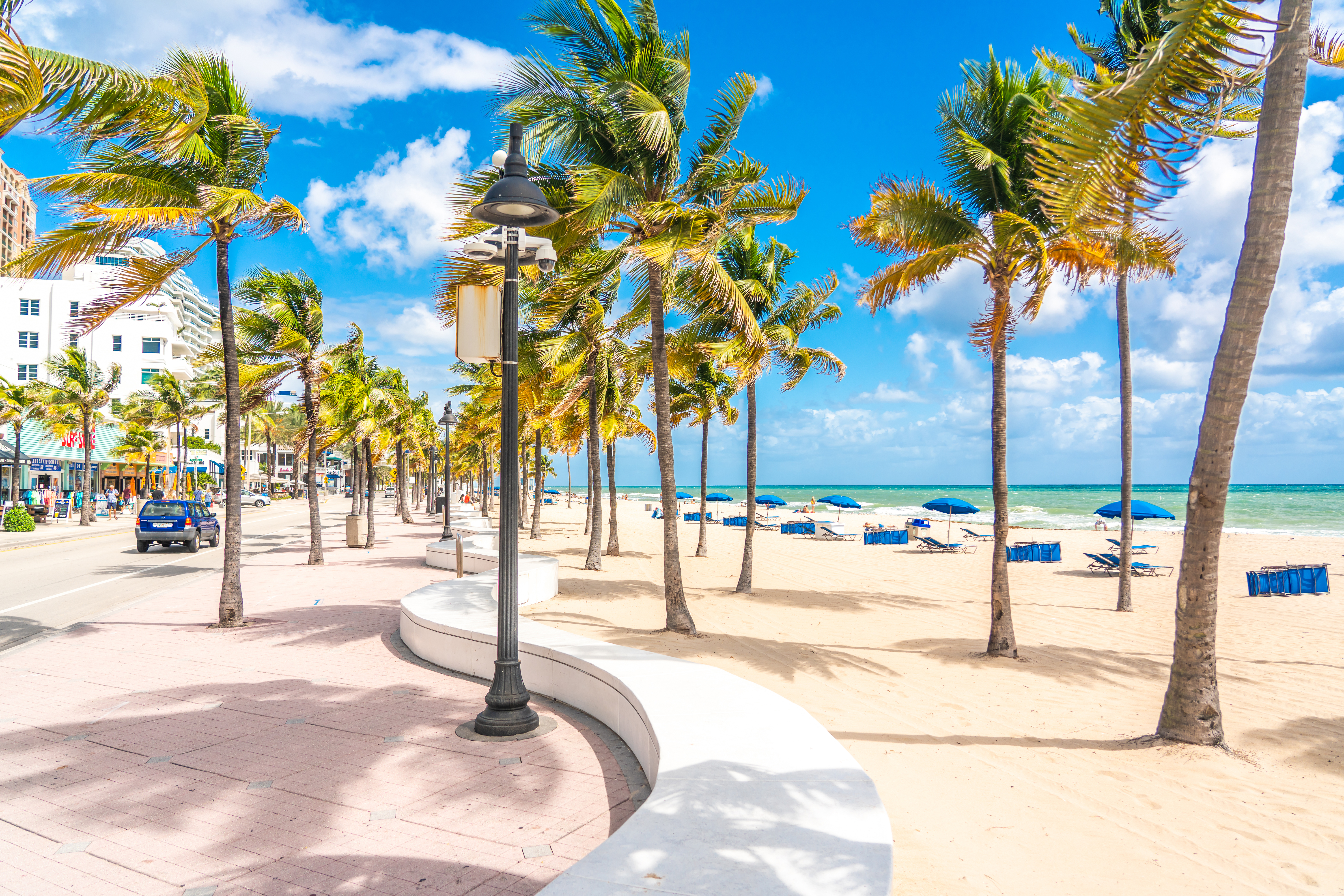 The seafront beach promenade in Fort Lauderdale is lined with palm trees, while blue sun loungers are laid out on the white-sand beach beyond. 